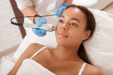 Happy beautiful African woman smiling while getting skin tightening treatment by beautician clipart