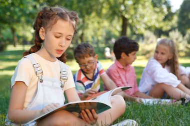 Parkta kitap okuyan genç bir kız. Arkadaşları arka plandaki çimlerde dinleniyor.