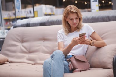 Akıllı telefonunu kullanan neşeli olgun bir kadın, mobilya dükkanında bir koltukta oturup, fotokopi çekiyor.