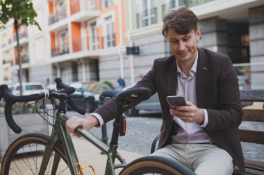 Akıllı telefon kullanan neşeli, olgun bir adam bisikletinin yanındaki bankta oturuyor.