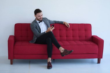 Genç bir adam oturuyor ve kırmızı kanepede bir akıllı telefon kullanma.