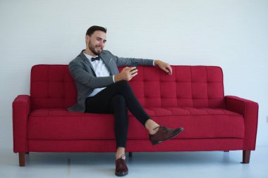 Genç bir adam oturuyor ve kırmızı kanepede bir akıllı telefon kullanma.
