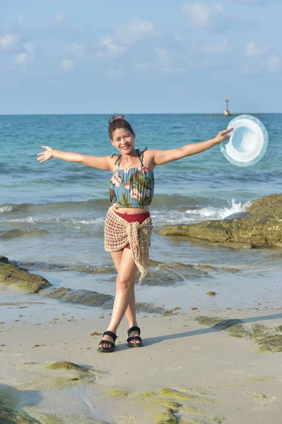 The beautiful girl is sending a sweet smile and a cheerful gesture with the view of the sandy beach, the blue sea, the waves and the sky on a bright day.