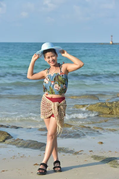 The beautiful girl is sending a sweet smile and a cheerful gesture with the view of the sandy beach, the blue sea, the waves and the sky on a bright day.