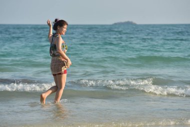 Güzel kız parlak bir günde kumlu plaj, mavi deniz, dalgalar ve gökyüzü manzarası ile tatlı bir gülümseme ve neşeli bir jest gönderiyor.