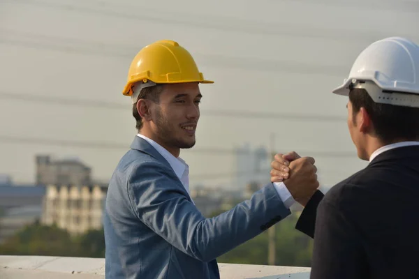 Businessmen in hard hats working together, shaking hands on city background.
