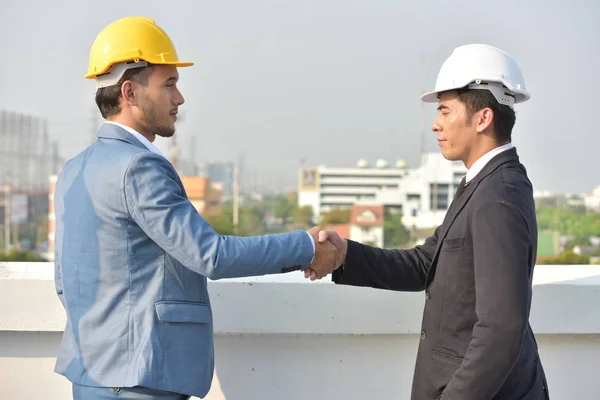 Businessmen in hard hats working together, shaking hands on city background.
