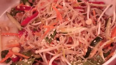 Çıtır havuç, salatalık, kırmızı çan biberi, fasulye filizi, çıtır fıstık ve susam tohumlarıyla süslenmiş cam şehriye salatası, ahşap yemek çubuklarının yanında beyaz seramik kasede dinleniyor.