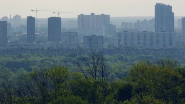 Kyiv kentsel ufuk çizgisi yemyeşil ormanın üzerinde görkemli bir şekilde yükseliyor, gündüz hava perspektifi sırasında metropol manzarası ile doğal ortam arasındaki uyumlu etkileşimi ortaya koyuyor.