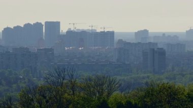 Kyiv 'in kentsel ufuk çizgisi, mevcut yapılar, ağaçlar ve puslu gökyüzü arasında yeni binalar üzerinde çalışan inşaat vinçleri gösteriyor