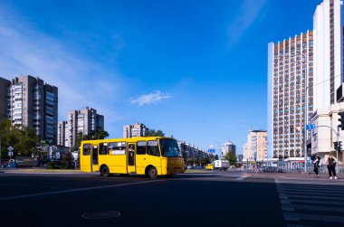 Kyiv, Ukraine 5th May,2025, Yellow bus drives through a street in Kyiv city surrounded by tall residential and office buildings, pedestrians crossing, and vibrant blue sky highlighting urban life