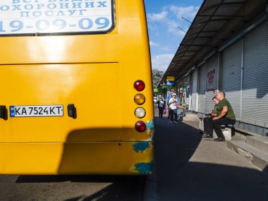 Kyiv, Ukraine 5th May,2025, Urban street scene in Kyiv with a yellow city bus at a bus stop, people waiting near closed market stalls, and sunlight casting shadows on the pavement