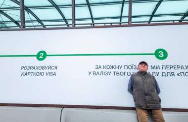 Kyiv, Ukraine 5th May,2025, Elderly man stands leaning casually against a wall with Ukrainian text beneath a glass canopy in Kyiv city, reflecting urban daily life, signage, and waiting concept