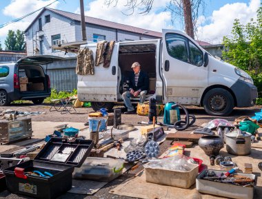 Kyiv, Ukraine 11th May,2025, Elderly man seated in open white van sells secondhand tools, hardware, and household items at outdoor flea market in residential area under bright summer sky