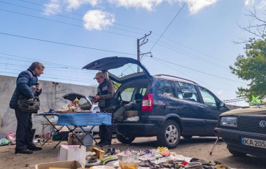 Kyiv, Ukraine 11th May,2025, Urban flea market setting where two men interact by a car with its trunk open, selling assorted second hand items on tables and ground with vehicles parked nearby