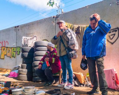 Kyiv, Ukraine 11th May,2025, Street market scene with men in casual clothing surrounded by stacked tires, used household goods, graffiti covered wall and sunlight casting urban atmosphere