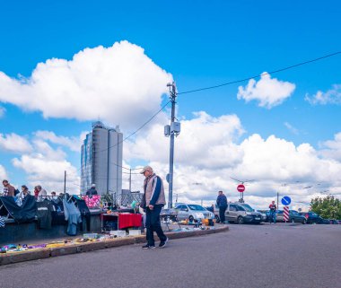 Kyiv, Ukraine 11th May,2025, Outdoor flea market scene with people walking past street stalls on a sunny day. Urban backdrop includes tall modern building, cars, and vivid sky with clouds