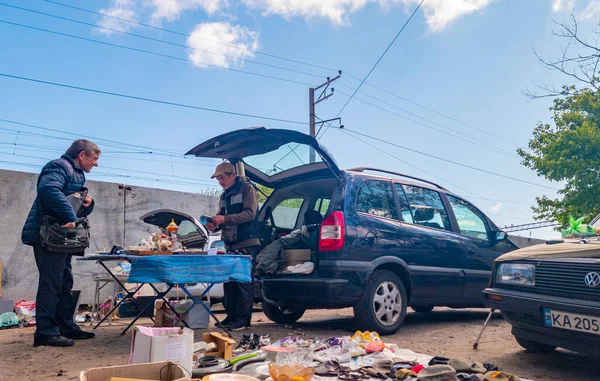 Kyiv, Ukraine 11th May,2025, Urban flea market setting where two men interact by a car with its trunk open, selling assorted second hand items on tables and ground with vehicles parked nearby