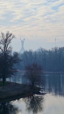 Hazy morning view of the Mother Ukraine monument, formerly known as the Motherland Monument, standing tall over the misty Dnipro River and a park with bare trees during a tranquil day in Kyiv