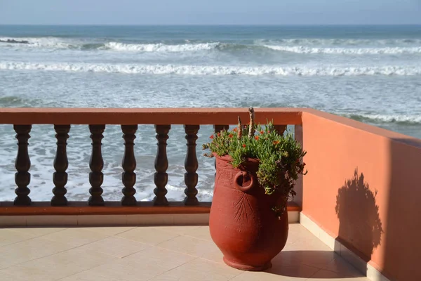 A large clay pot with a plant stands on a terrace overlooking the Atlantic Ocean on a sunny day.