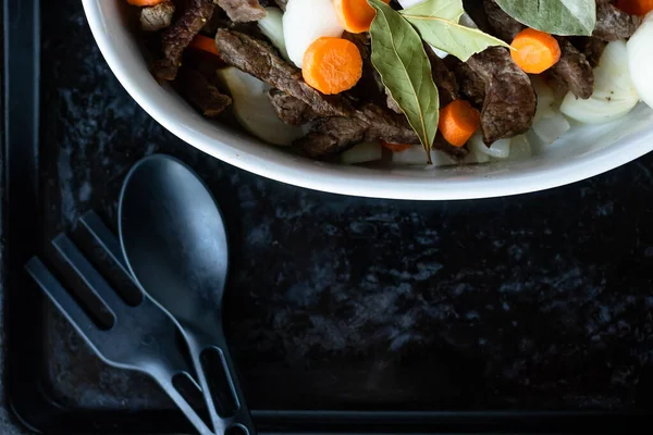 Siyah tepside et, havuç, soğan ve defne ile beyaz tabak. Yemek malzemeleri kaşığı ve çatalı. Üst Manzara. Boşluğu kopyala.