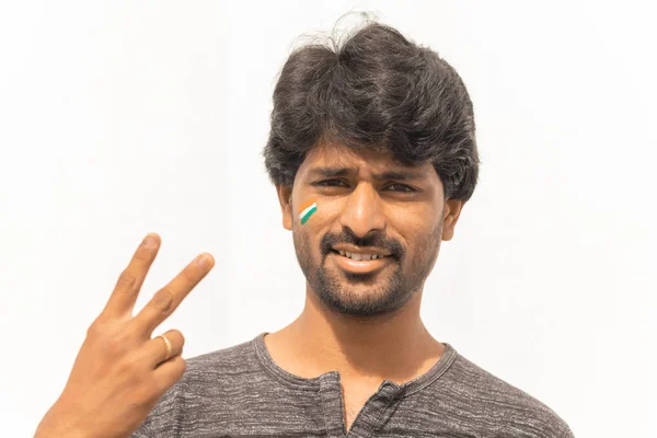 Portrait de jeune homme indien joyeux fan de sport de cricket Drapeau indien peint sur son visage et montrant le geste de la main de la victoire — Photo