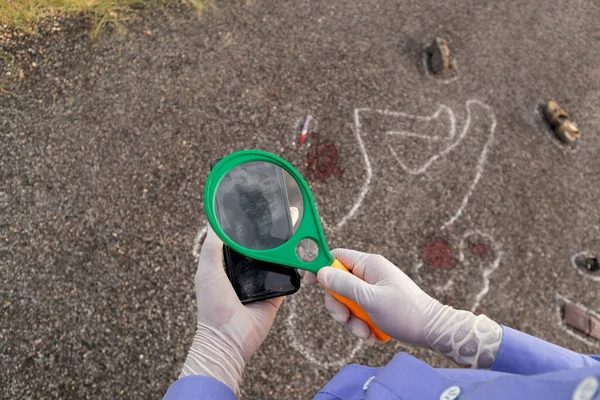 Dedektif ya da araştırmacı cep telefonundaki parmak izlerini doğrulamak için büyüteç ve tebeşir hatlarını arka plan olarak kullanıyor.