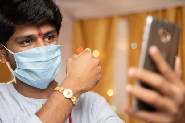 Homem em máscara médica ocupado no telefone celular e mostrando Rakhi ou RakshaBandhan para irmã ou amigos da família na cerimônia do festival durante coronavírus ou covid-19 pandemia em casa com luzes de decoração . — Fotografia de Stock