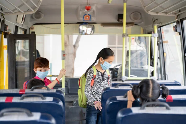 Kids with medical mask coming inside school bus and sitting on seats while maintaining social distance due to coronavirus or covid-19 pandemic - Concept of school reopen or back to school