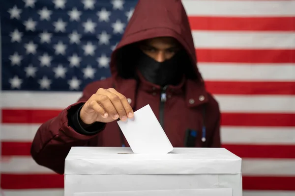 Людина в худі з покритим обличчям лиття Голосувати на виборчій кабінці з US falg як тло - Концепція невідомого голосування або фальсифікації голосів на виборах США. — стокове фото