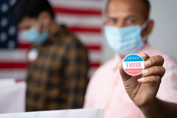 Закриття Руків'я, Людина в медичній масці показує, що я голосував Sticker на виборчій кабінці з прапором США як тло - концепція в голосуванні людини на виборах США. — стокове фото
