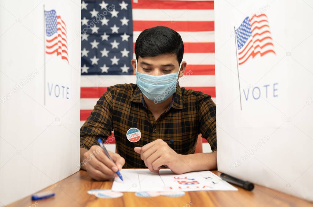 Young man in medical mask busy inside the polling booth with US flag as background - Concept of in person voting with covid-19 safety measure at US election.