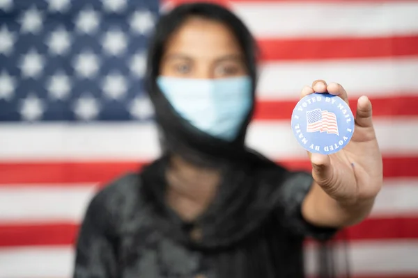 Foco seletivo em mãos, Menina com Hijab ou cobertura da cabeça e máscara usado mostrando Eu votei Adesivo com bandeira dos EUA como fundo Conceito de votação durante a eleição dos EUA. — Fotografia de Stock
