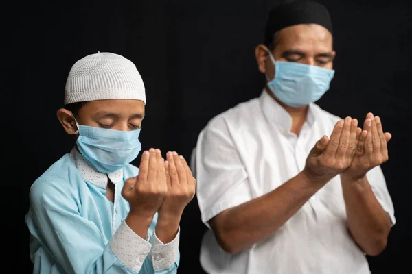 Pai muçulmano e filho em máscara médica orando ou realizando Salah o proteger do coronavírus ou covid-19 sentando. — Fotografia de Stock