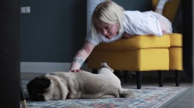Genç bir kadın köpeğini sandalyede otururken okşadı. Yerde uyuyan sevimli köpek yavrusu. Aşk ve özen sahnesi.