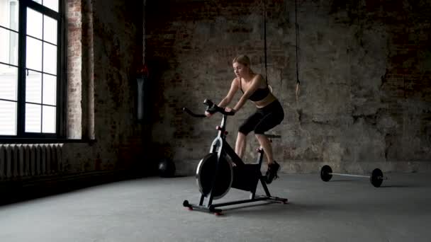 可爱的女孩化妆和发型做健身在一个固定的自行车在黑暗的健身房 在框架腿 慢动作 — 图库视频影像