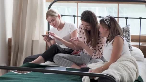Három lány ül és csevegnek egy ágyon egy szobában, képeket néznek okostelefonokon és mosolyognak.