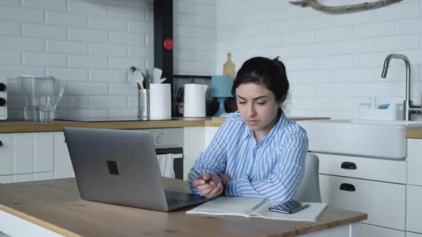 一位身穿蓝色条纹衬衫的印度女孩在家里的电脑上工作 并在明亮厨房的笔记本上记笔记 — 图库视频影像