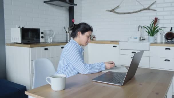 在厨房里 一位严肃的印度妇女坐在桌旁 坐在电脑前 一边喝咖啡 一边认真地做着在线工作 — 图库视频影像