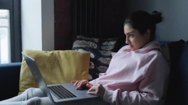 Genç Hintli kadın evdeki koltukta oturuyor ve bilgisayar üzerinde çalışıyor.