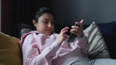 Oturma odasındaki kanepede uzanmış rahat genç bir kadın akıllı telefon kullanıyor. Tembel mutlu milenyum bayanı sosyal medyada sörf yapıyor, uygulama alışverişi yapıyor, mobil oyun oynuyor, cep telefonuna mesaj yazıyor..