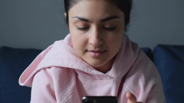 スマートフォンをオンラインで使用し ソファの上に自宅で座っている若いインド人女性の肖像画を閉じます — ストック動画