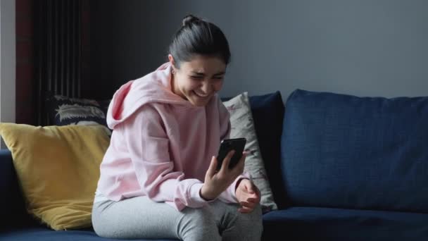 若いインド人の女性は 自宅でソファの上で笑顔と笑って スマートフォン上でテキストメッセージ — ストック動画