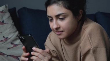 Sırıtkan genç bir Hintli kadının yatakta uzanmış akıllı bir telefonla haberleri okuması ve sosyal medyayı izlemesi.
