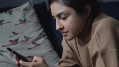 Yatağa uzanmış, akıllı bir telefonla haberleri okuyan ve sosyal medyayı izleyen genç bir Hintli kadının yan görüntüsü.