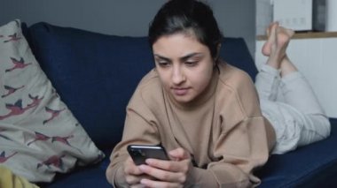  Genç Hintli bir kadın yatakta uzanmış, akıllı bir telefonla haberleri okuyor ve sosyal medyayı izliyor.