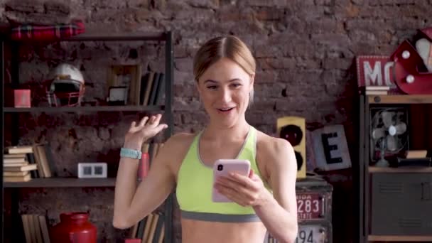 在一个装有大窗户的健身工作室里 一个年轻的金发女人用智能手机在网上聊天 — 图库视频影像