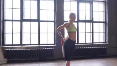 Genç, atletik sarışın bir kadın büyük pencereli bir spor salonunda ısınma egzersizi yapıyor.