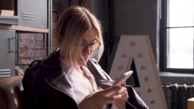 Gözlüklü, sarışın genç bir iş kadını iş yerinde akıllı bir telefon kullanır ve büyük, parlak bir ofiste bir sandalyede oturur.