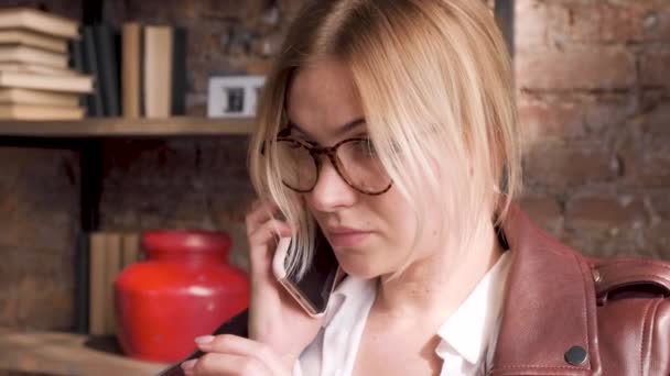 在一个明亮的大办公室里 一个戴着眼镜的年轻女商人用智能手机俯视下面 坐在椅子上讲电话的画像 — 图库视频影像